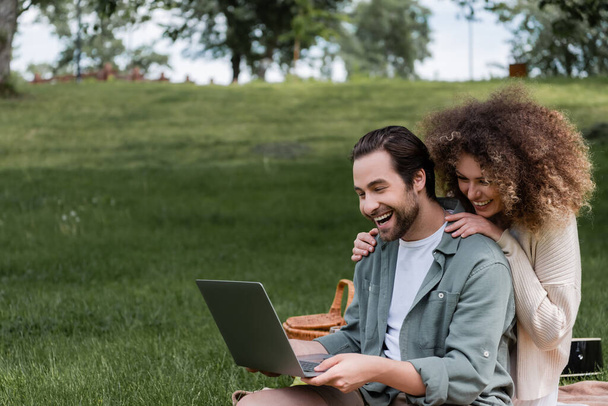 изумленный мужчина с ноутбуком рядом счастливая женщина во время пикника в парке - Фото, изображение