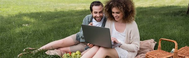 ευτυχισμένη γυναίκα που χρησιμοποιεί φορητό υπολογιστή, ενώ κάθεται σε κουβέρτα με το φίλο κατά τη διάρκεια πικνίκ, πανό - Φωτογραφία, εικόνα