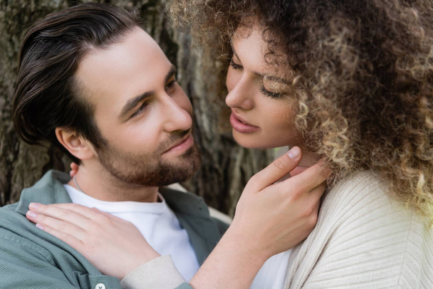 portrait of young and happy man touching face of curly woman near tree trunk  - Foto, Bild