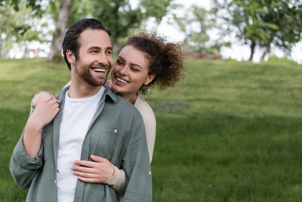 pleased and curly woman hugging boyfriend from behind in green summer park - Fotó, kép