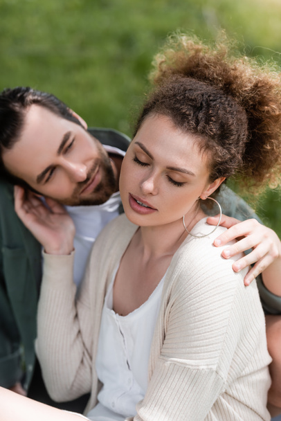 tender and curly woman touching face of bearded boyfriend in park - Fotoğraf, Görsel