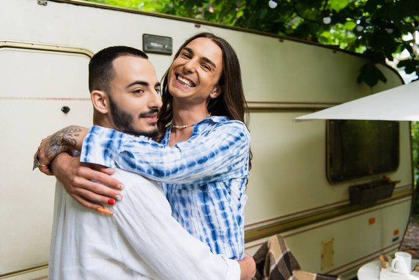 happy tattooed gay man hugging cheerful boyfriend near travel van  - Photo, image