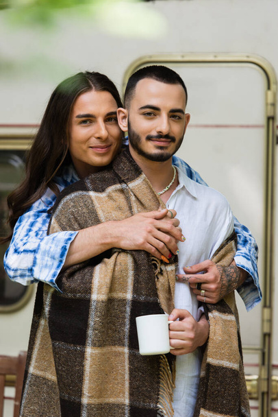 smiling man with tattoo hugging bearded boyfriend in blanket near blurred van - Valokuva, kuva