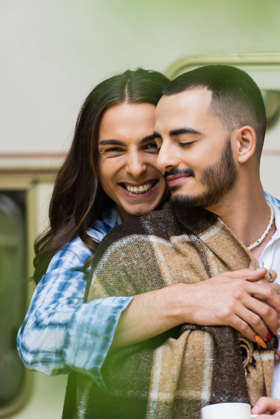 joyful man hugging bearded boyfriend in blanket near blurred van - Foto, immagini