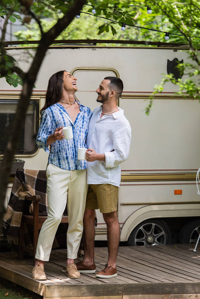 full length of happy gay couple holding cups of coffee while laughing near van in summer - Photo, image
