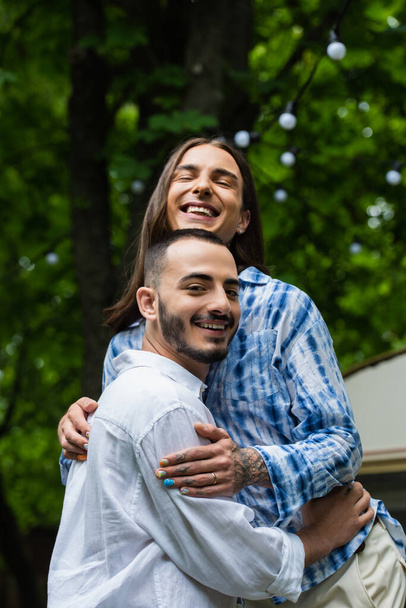 happy married gay couple smiling while hugging near blurred travel van  - Foto, Bild