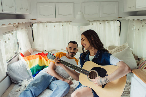 счастливый и татуированный гей, играющий на акустической гитаре рядом с парнем на кровати в современном фургоне  - Фото, изображение