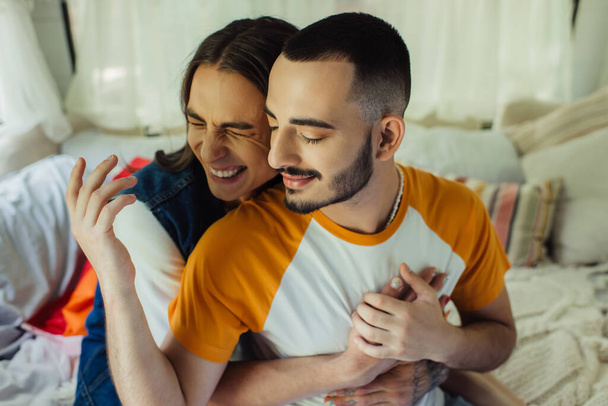 high angle view of smiling gay man with tattoo hugging bearded boyfriend with closed eyes in modern van  - Fotoğraf, Görsel