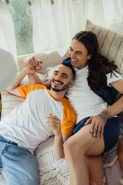 high angle view of smiling gay man with tattoos lying on bed with bearded boyfriend in modern van  - Zdjęcie, obraz