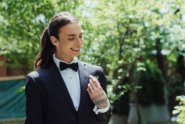 happy tattooed man with golden wedding ring on finger smiling while adjusting floral boutonniere  - 写真・画像