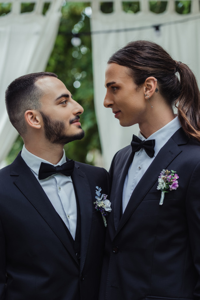 side view of gay couple in suits with boutonnieres looking at each other during wedding ceremony  - Foto, afbeelding