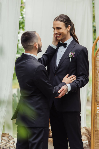 bearded gay man touching cheek of pleased groom on wedding day - Photo, image