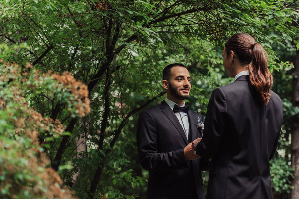happy gay newlyweds in formal wear looking at each other and holding hands in park - Photo, Image