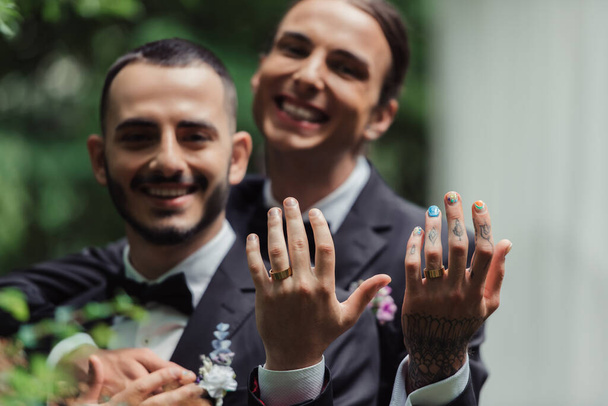 blurred and happy gay newlyweds in formal wear with boutonnieres showing golden rings on fingers  - Photo, Image