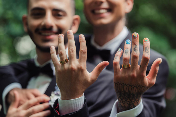 blurred and cheerful gay newlyweds in formal wear with boutonnieres showing golden rings on fingers  - Zdjęcie, obraz