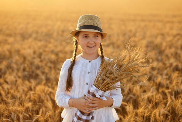 Niña en un campo de trigo. Una chica con sombrero de paja. Niña sosteniendo espigas de maíz en sus manos. Imagen con enfoque selectivo. - Foto, imagen