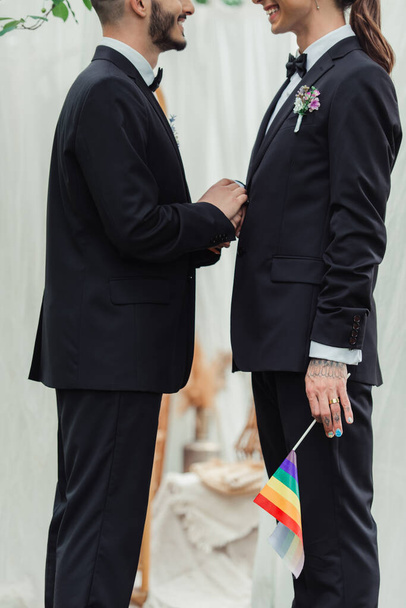 Ausgeschnittene Ansicht von glücklichen homosexuellen Brautpaaren in formeller Kleidung mit lgbt-Fahne während der Trauung  - Foto, Bild