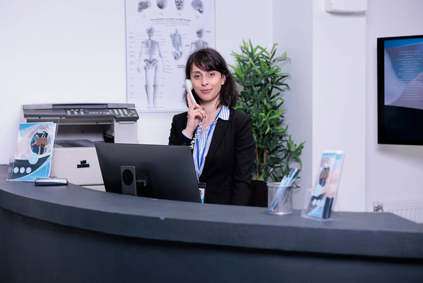 Portrait of smiling hospital receptionist answering call from patient to make an appointment for clinical consult at private clinic. Professional looking woman working at front desk using telephone. - Photo, Image