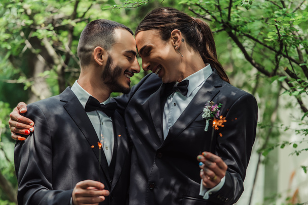 smiling gay couple in formal wear holding sparklers in green park on wedding day - Photo, Image