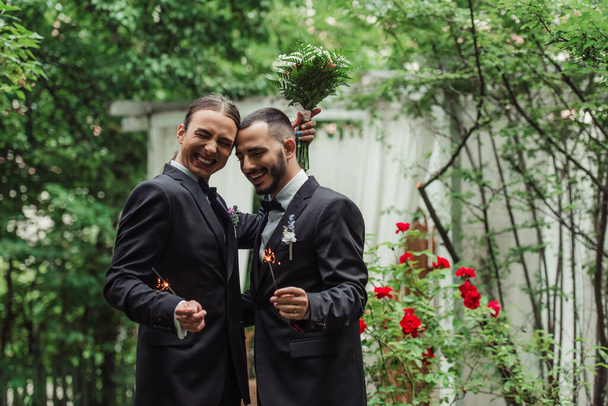 happy gay man in suit holding wedding bouquet and sparkler near groom in green park - Fotoğraf, Görsel