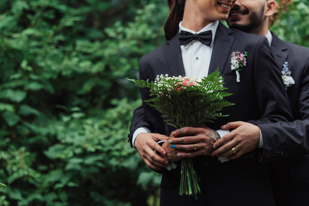cropped view of joyful gay couple in suits holding wedding bouquet in green park - Fotoğraf, Görsel