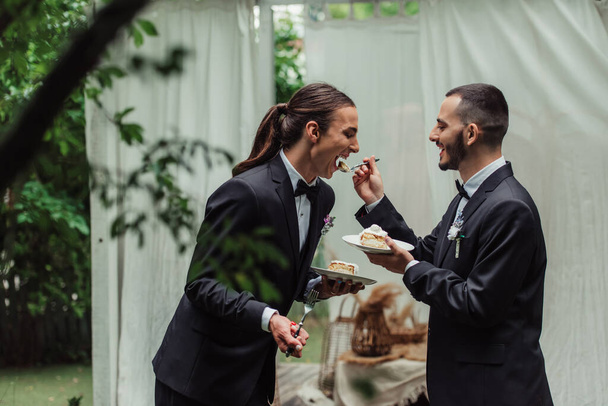 happy gay man in formal wear feeding husband with wedding cake  - Foto, Imagen