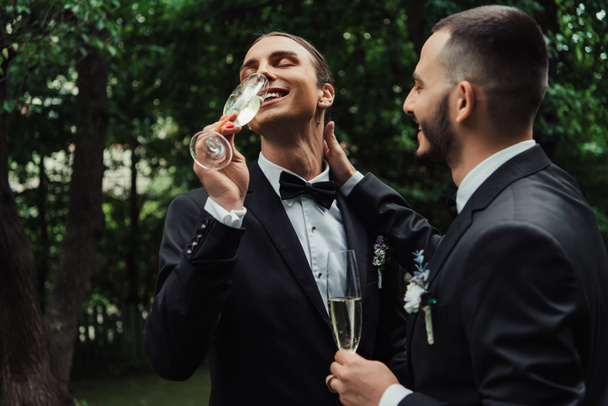 happy gay newlyweds in suits drinking champagne on wedding day  - Photo, Image