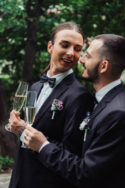 happy gay newlyweds in suits holding glasses with champagne and looking at each other - Fotoğraf, Görsel
