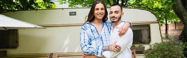 happy gay couple smiling while hugging near travel van in forest, banner - 写真・画像