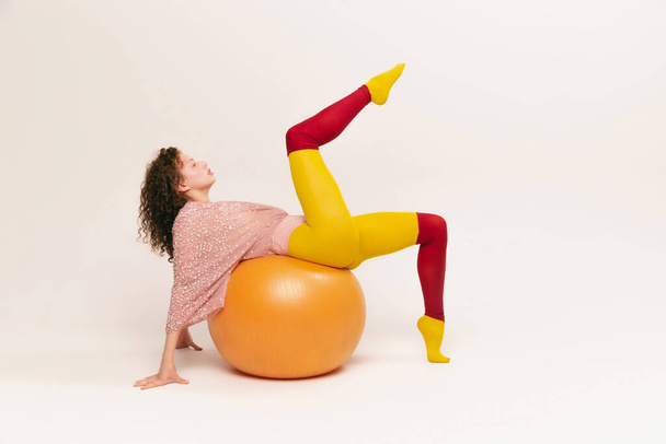Doing exercises on ball. Young pretty funny girl in avant-garde style clothes having fun over white background. Fitness, vivid style, beauty, queer, freak, fashion concept. - Photo, image