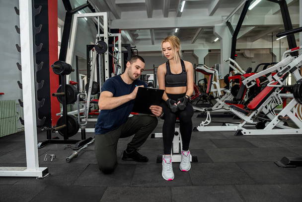 Fit female client working out with a dumbbell under control of the personal male trainer at modern gym. Sport as lifestyle - Zdjęcie, obraz
