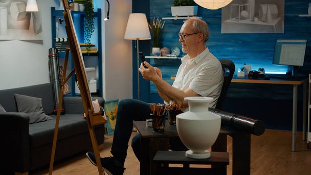 Üst düzey bir adam vazo başyapıtında cep telefonuyla fotoğraf çekiyor, sanatsal ilham bulmak için akıllı telefonu kullanıyor. Evde sanat ve zanaat hobisi için çizim ve çizim yapıyorum.. - Fotoğraf, Görsel