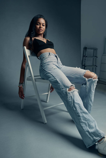 Young Latin American woman in stylish jeans and bra relaxing on chair and looking at camera in studio with gray walls - Photo, Image