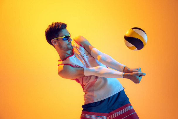 Portrait of young man, volleyball player in motion, bumping ball, playing isolated over yellow studio background in neon light. Concept of sport, action, team game, active lifestyle, health, hobby, ad - 写真・画像