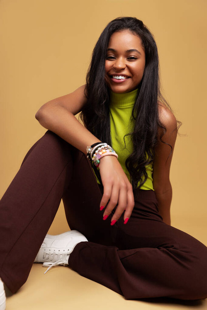 Cheerful young Latin American female millennial with long dark hair in trendy outfit smiling, happily and looking at camera while sitting against beige background in studio - Zdjęcie, obraz