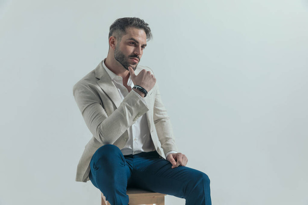 attractive young businessman in suit with grey hair touching chin and looking to side while holding elbow on knee and sitting on wooden chair in front of grey background in studio - Photo, Image