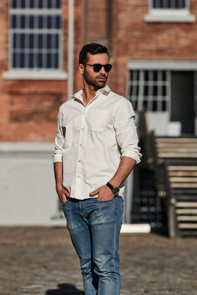 Self assured ethnic male in jeans and white shirt with stylish sunglasses holding hands in pockets and looking away, while standing on sunlit city street outside brick building - Photo, Image
