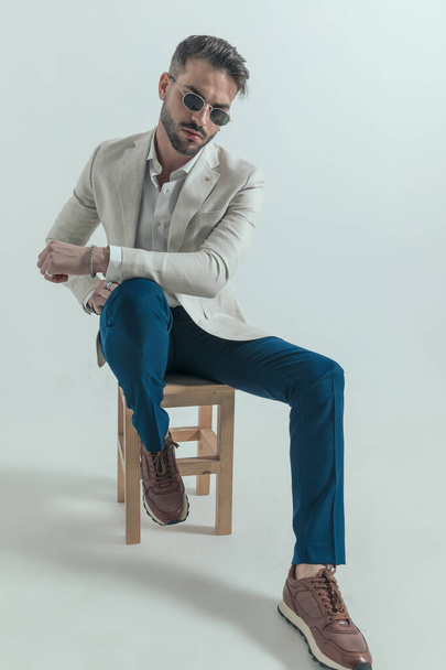 jovem atraente de terno com camisa desfeita segurando braços em uma pose de moda enquanto sentado em cadeira de madeira em fundo cinza no estúdio - Foto, Imagem