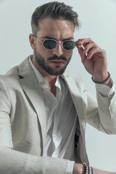 уверенный бизнесмен в костюме с расстегнутой рубашкой, регулирующий солнцезащитные очки и позирующий на сером фоне - Фото, изображение