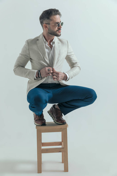 crouched bearded man looking to side and buttoning jacket while posing in front of grey background - Foto, Bild