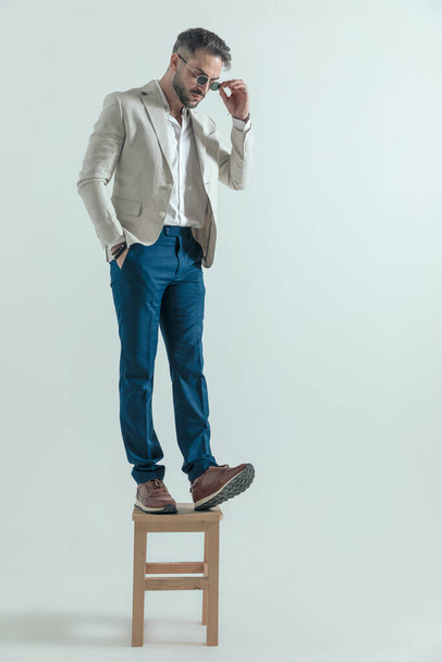 atractivo hombre en traje casual inteligente mirando hacia abajo en sus zapatillas de deporte y la fijación de gafas de sol, mientras que de pie en una silla de madera delante de fondo gris - Foto, Imagen