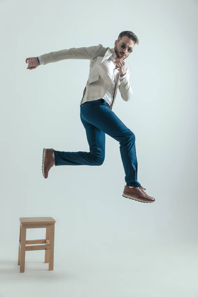 full body picture of bearded businessman with glasses jumping off wooden chair, touching chin and thinking about his leaving decision and new life on grey background - 写真・画像