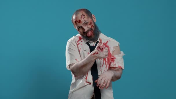Creepy looking zombie showing thumbs up hand gesture on blue background. Approving spooky brain-eating evil walking dead corpse with deep and bloody wounds showing approval hand sign. Studio shot - Footage, Video