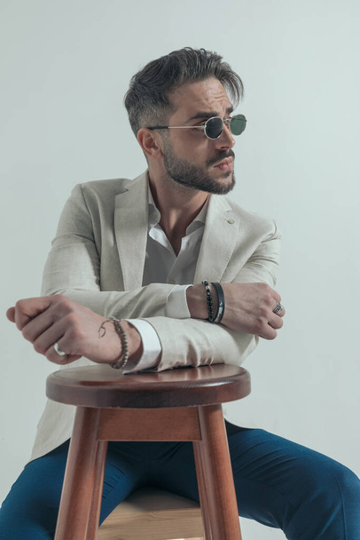 hombre de negocios barbudo mirando hacia un lado y sosteniendo los brazos en una silla de madera en una pose de moda sobre fondo gris sentado en el estudio - Foto, Imagen