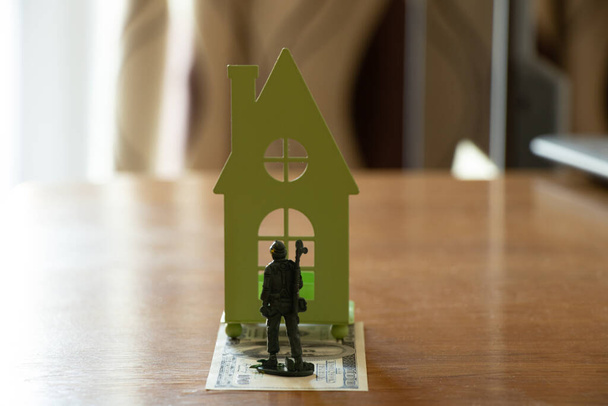 Soldat en plastique pour enfants debout sur les dollars, prendre une petite maison verte sur la table à l'appartement, l'achat d'une maison pour un soldat militaire, le succès et les finances, l'armée - Photo, image