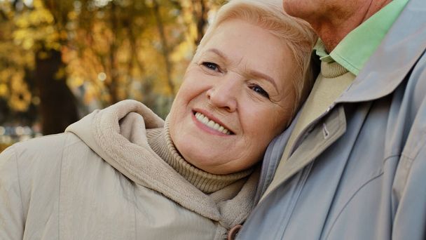 Happy middle aged pretty caucasian elderly woman grandma smiling stand relax in autumn park spend time outdoors lean on shoulder mature partner man supporting husband show empathy care love romantic - Photo, Image