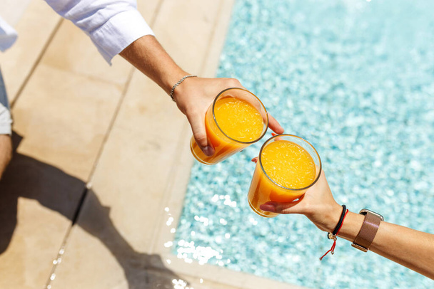 Top vista mãos de casal em desgaste casual sentado junto à piscina e beber coquetéis ou suco de laranja. Relaxe despreocupado na piscina. Conceito de vocação. - Foto, Imagem