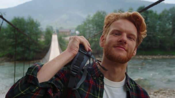 笑顔ハイカー川の橋の上にカメラをポーズ。幸せな旅行者の肖像はハイキングでバックパックを着用する。喜びの男は山の自然で観光レジャーをお楽しみください。ジンジャー男は屋外で見て回る。旅行のコンセプト. - 映像、動画