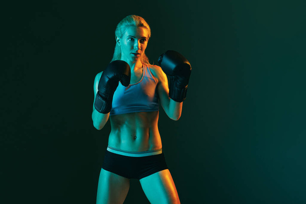 Punch. One young woman, female professional kickboxer in sports uniform and gloves training isolated on dark green background in neon. Sport, competition, power concept. - 写真・画像