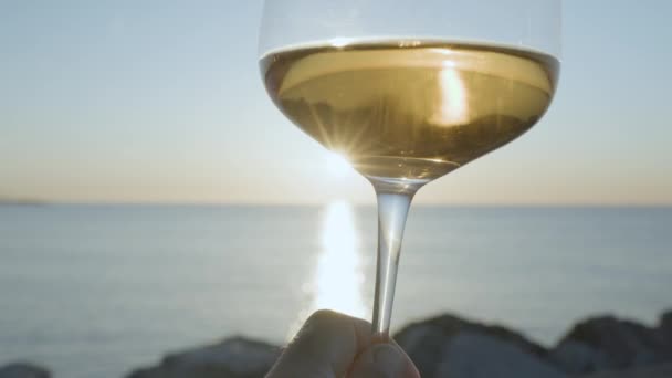 Sklenice na víno s bílým vínem se šumivým mořem a oblohou v pozadí při východu nebo západu slunce - Záběry, video